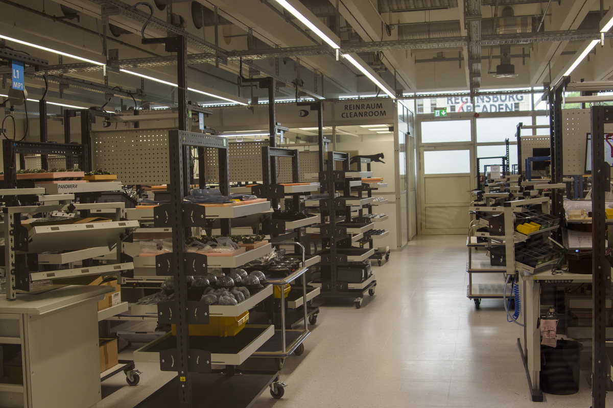 A spacious cleanroom environment filled with metal shelving units, organized with various equipment and devices, illuminated by overhead lights, with a door visible in the background.