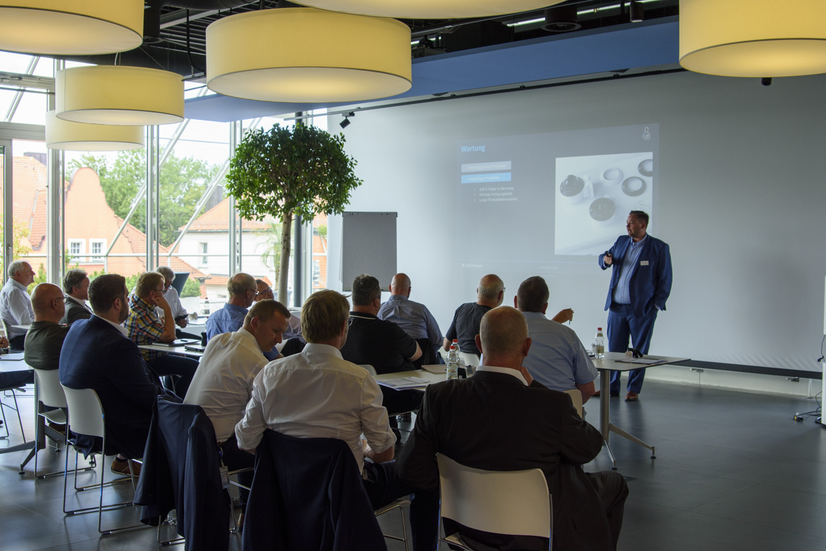A speaker presents to an audience in a modern conference room, with attendees seated at tables. A large screen displays a presentation.