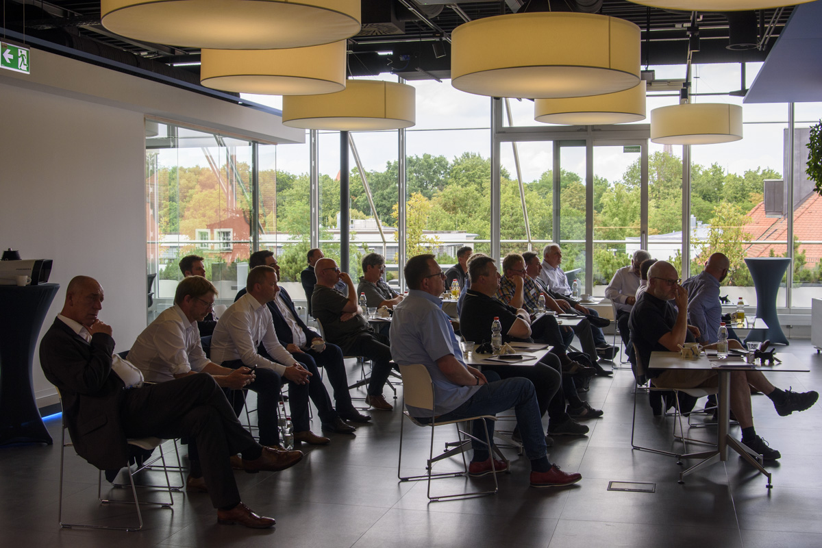 A group of people seated in an indoor conference setting, listening attentively, with large windows showing trees and buildings outside.