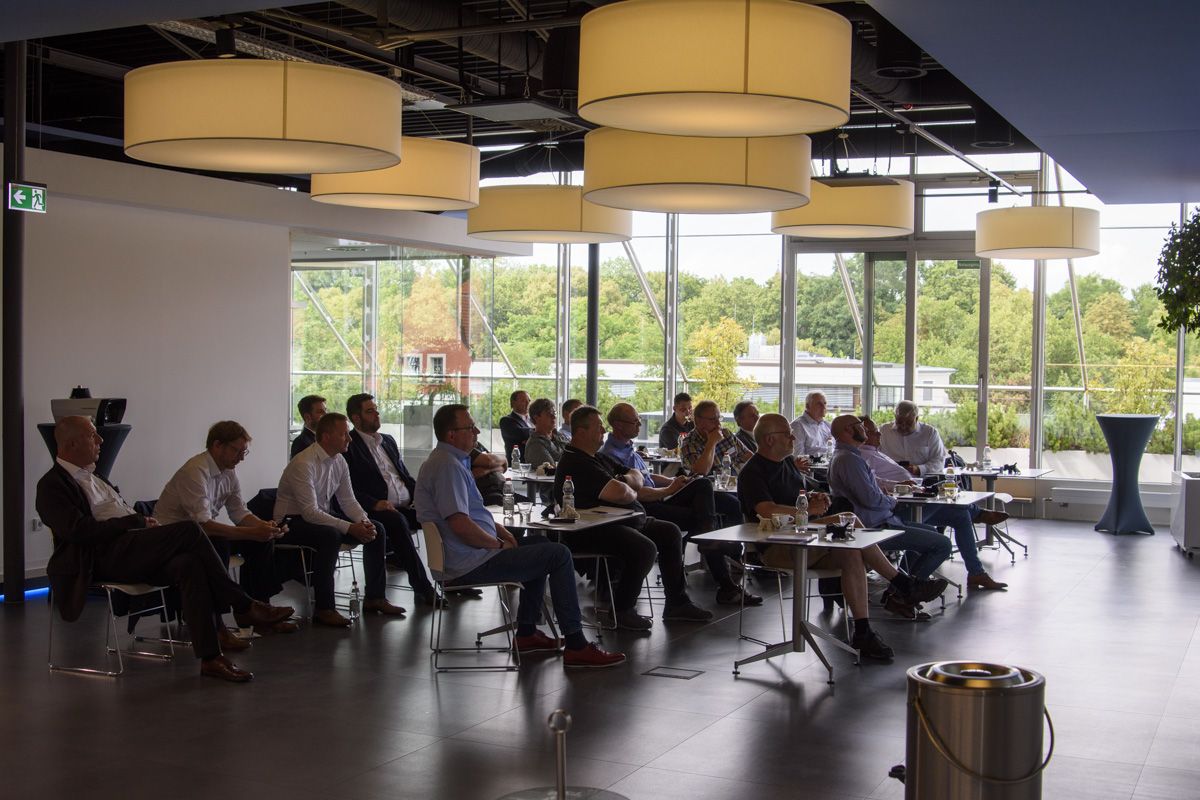 A group of people seated in a modern conference room, attentively listening to a presentation. Large windows with natural light and greenery visible outside.