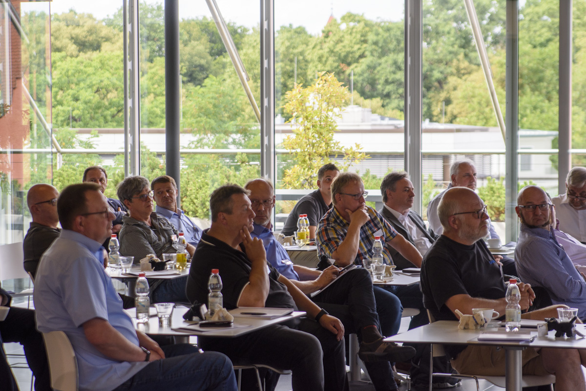 A group of men sitting at tables, attentively listening during a presentation or meeting in a modern space with large windows and greenery outside.