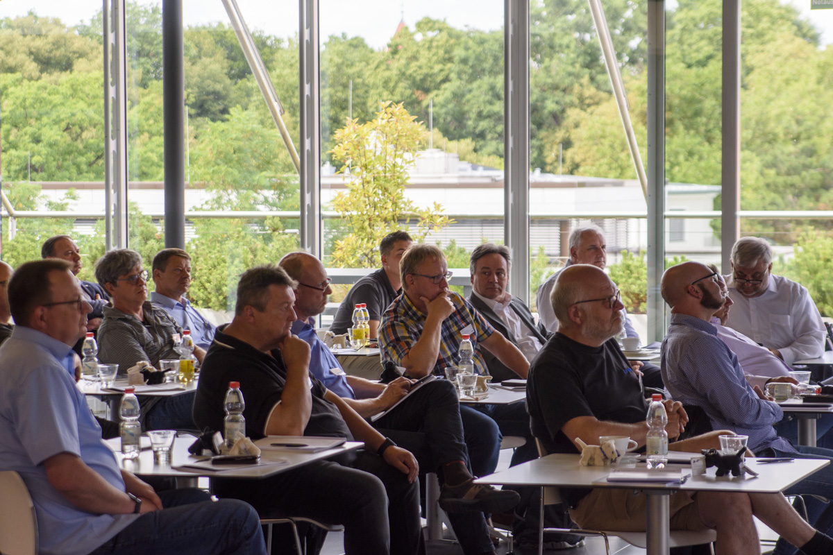 A group of people seated at tables in a modern room, attentively listening to a presentation, with greenery visible through large windows.
