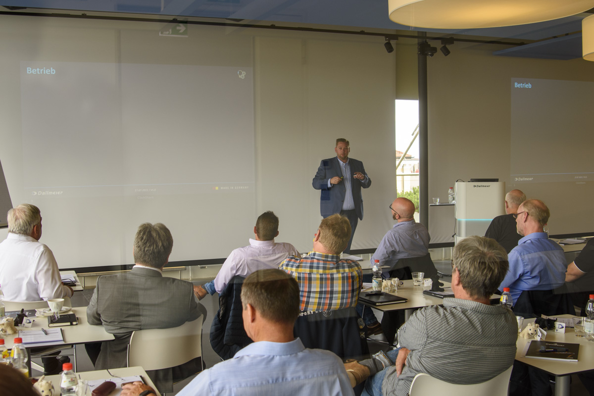 A presenter speaks to an engaged audience in a conference room, with attendees seated at tables. Large screens display a presentation titled "Betrieb.
