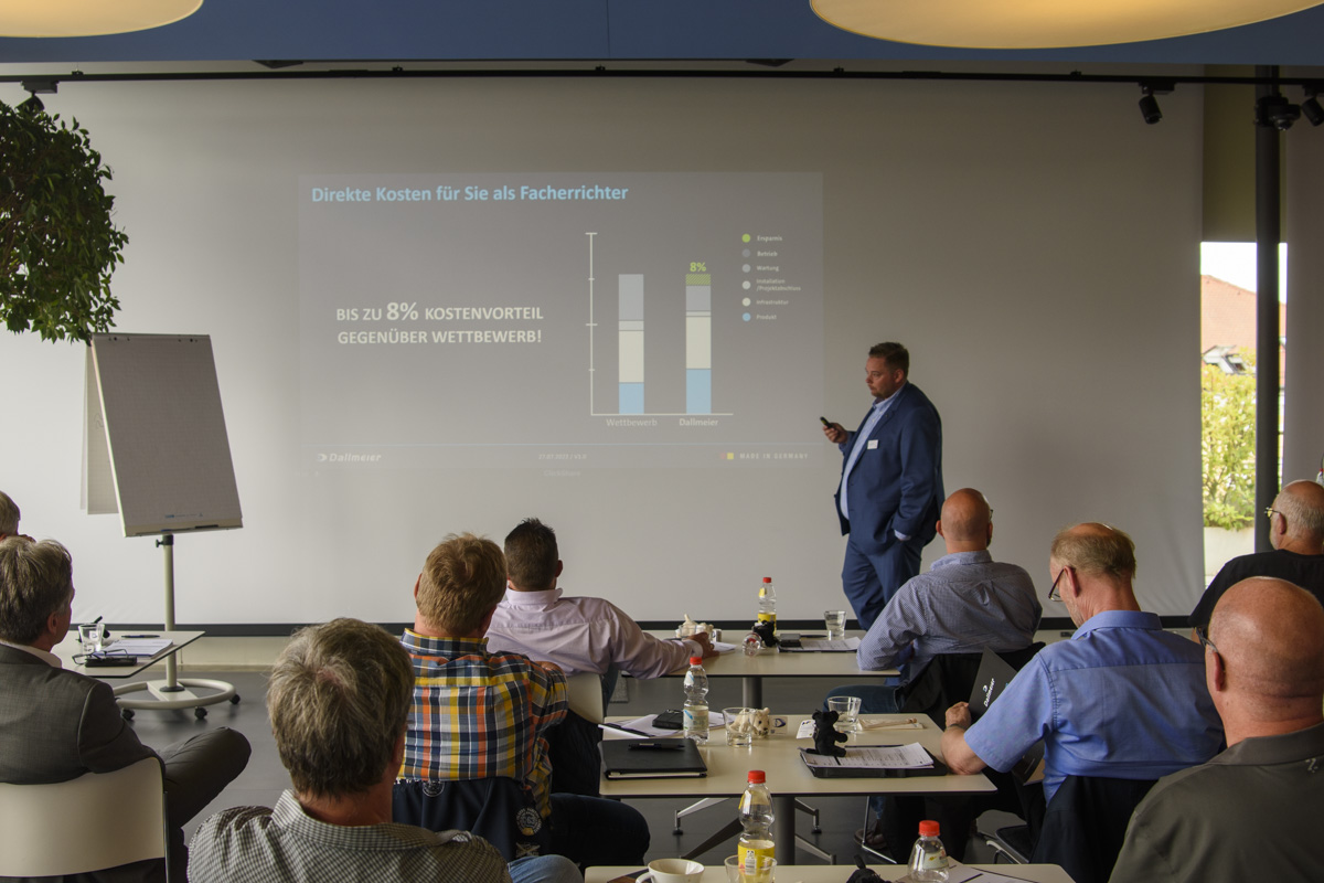 A presenter speaks to an audience in a meeting room, showcasing a graph on a screen about cost comparisons in a business context. Attendees are seated at tables.