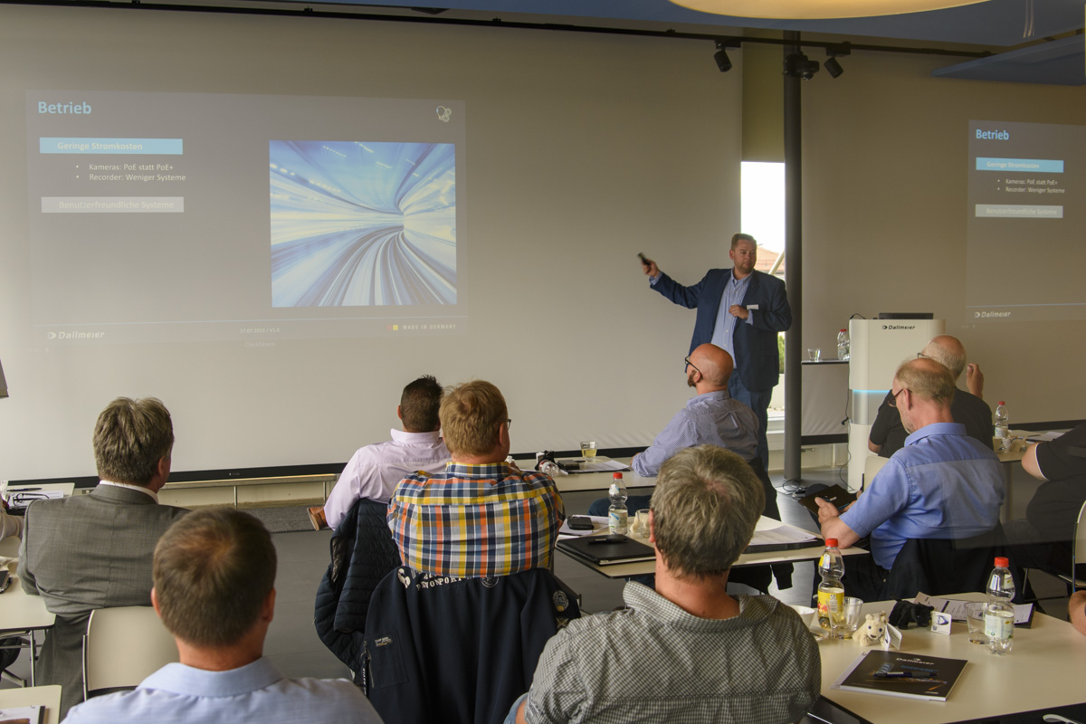 A presenter stands in front of a screen displaying graphics, addressing an audience seated at tables during a conference or meeting.
