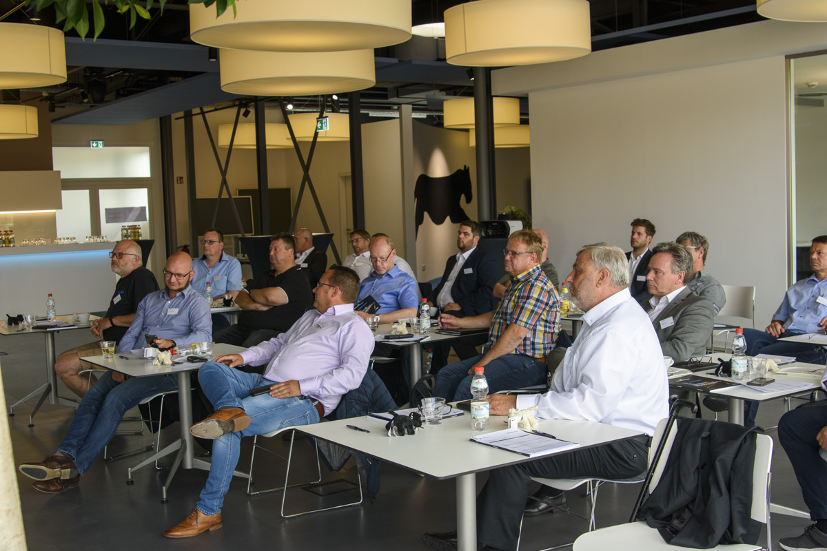A group of people seated at tables in a conference room, attentively listening to a presentation. The room is well-lit with modern decor and overhead lighting.