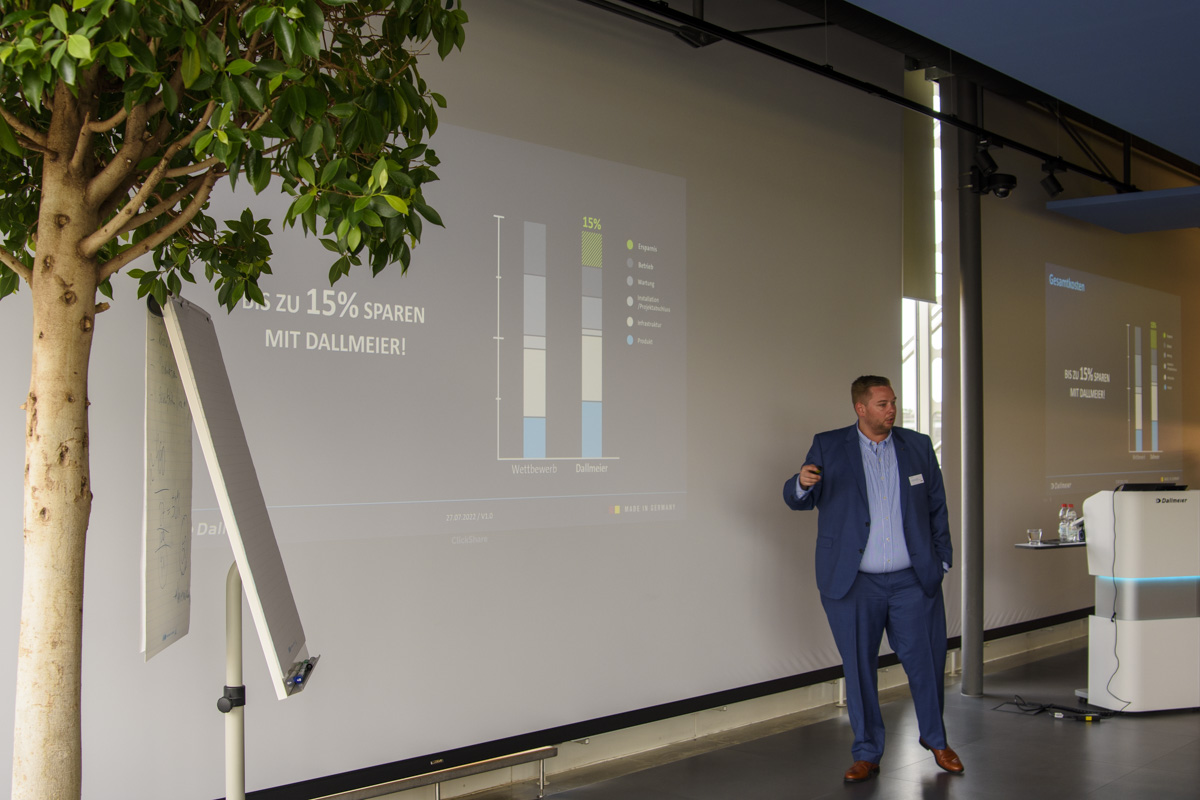 A man in a suit presents data on a large screen, indicating potential savings of up to 15%. A potted plant is visible beside him.