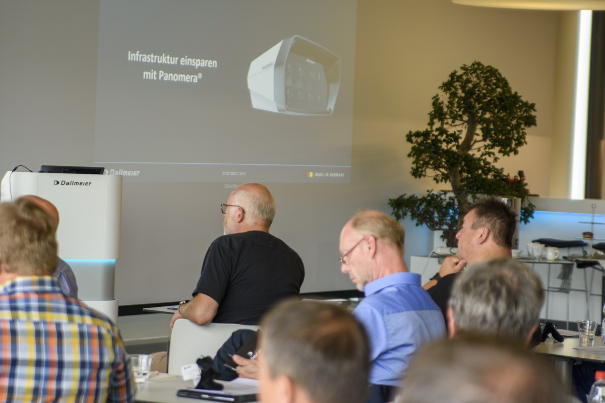 A group of people seated at a conference room table, focusing on a presentation screen displaying the text "Infrastruktur einsparen mit Panomera" alongside an image of a device.