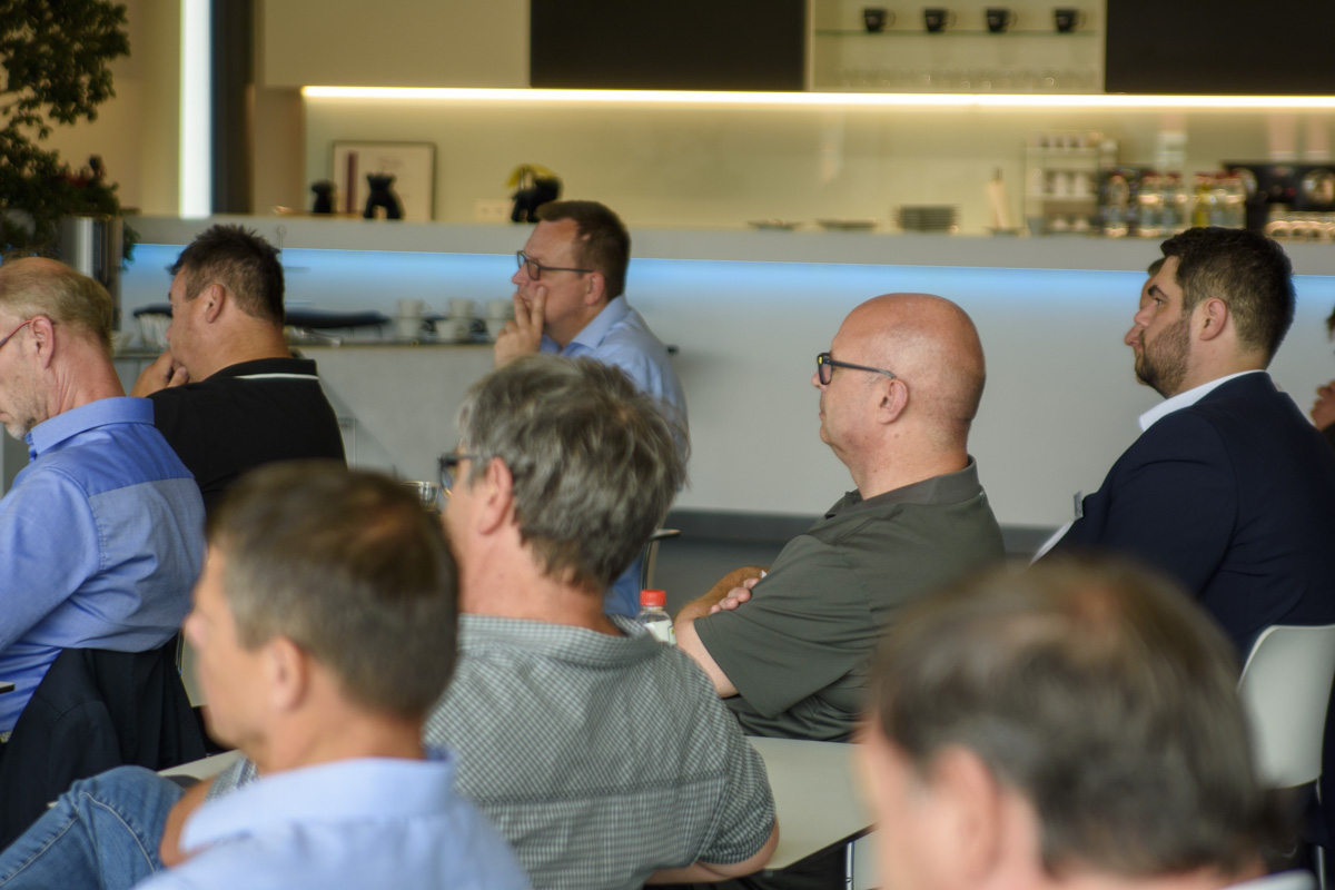 A group of people seated in a modern setting, attentively listening, with a bar area visible in the background.