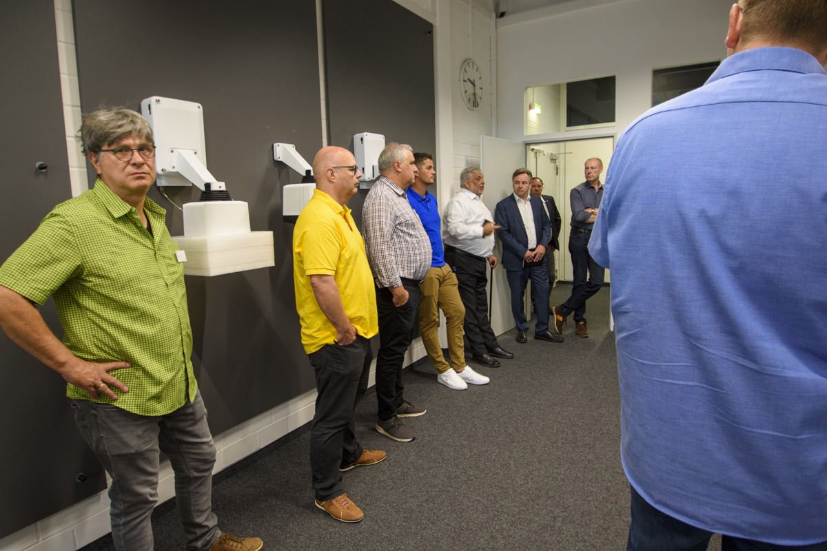 A group of men stands in a room, engaged in conversation, with some wearing brightly colored shirts. Wall-mounted devices are visible in the background.