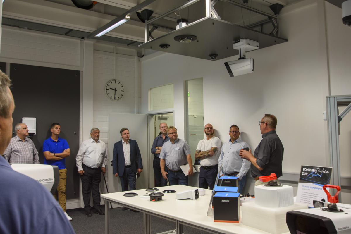 A group of people listens to a speaker in a modern presentation room, featuring tables with technology, a clock on the wall, and a projector above.