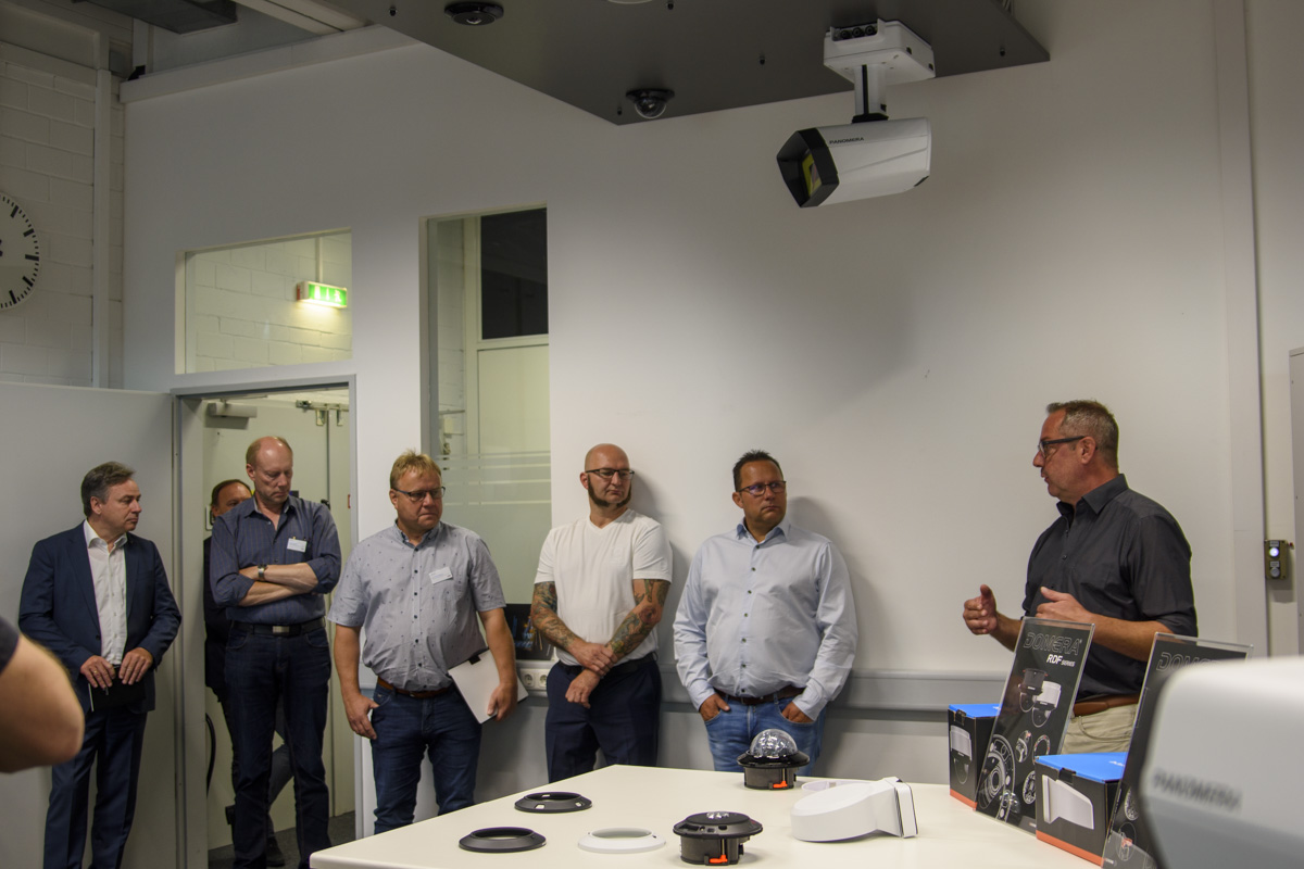 A group of six people in a room, listening to a speaker who stands near a table with various equipment. A projector hangs from the ceiling.