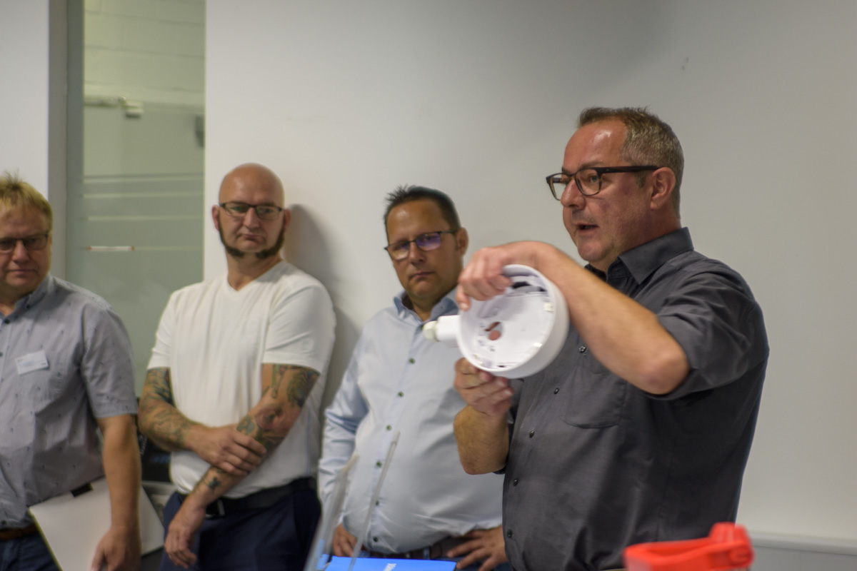 A man in a gray shirt holds a round object while explaining to a group of three men. They are engaged in a discussion in a well-lit room.