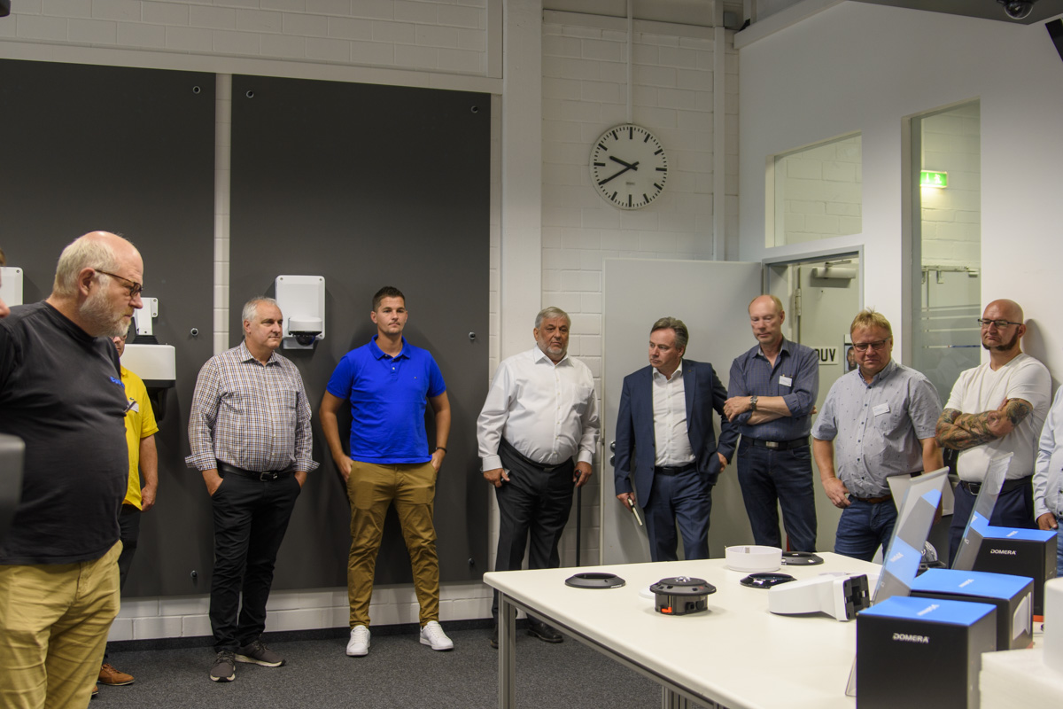 A group of men are gathered in a room, listening intently to a presentation near a table displaying various tech devices. A clock is visible on the wall.