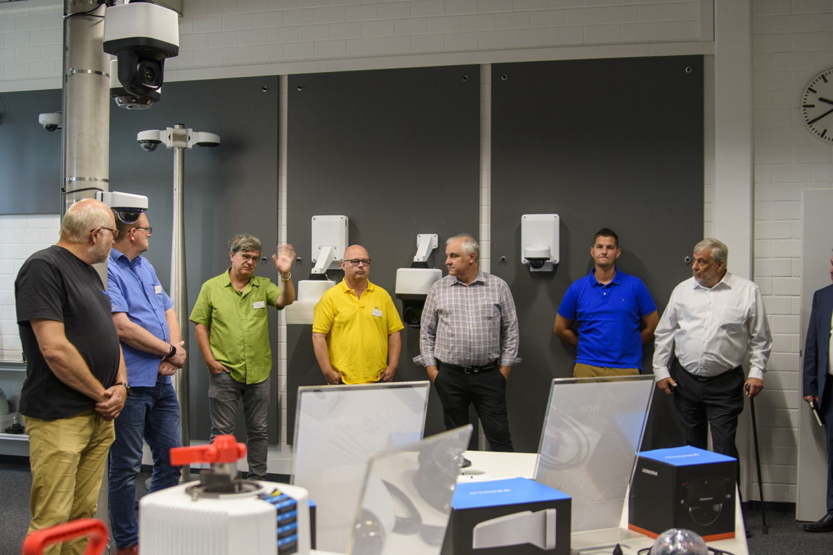 A group of eight men stands in a workshop, engaged in discussion, with various equipment and tools displayed on a table in the foreground.