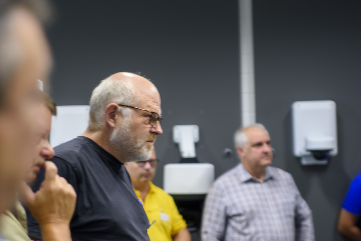 A group of people is gathered in a room, focused on a discussion. One man with glasses and a bald head appears contemplative. Various handwashing stations are visible in the background.
