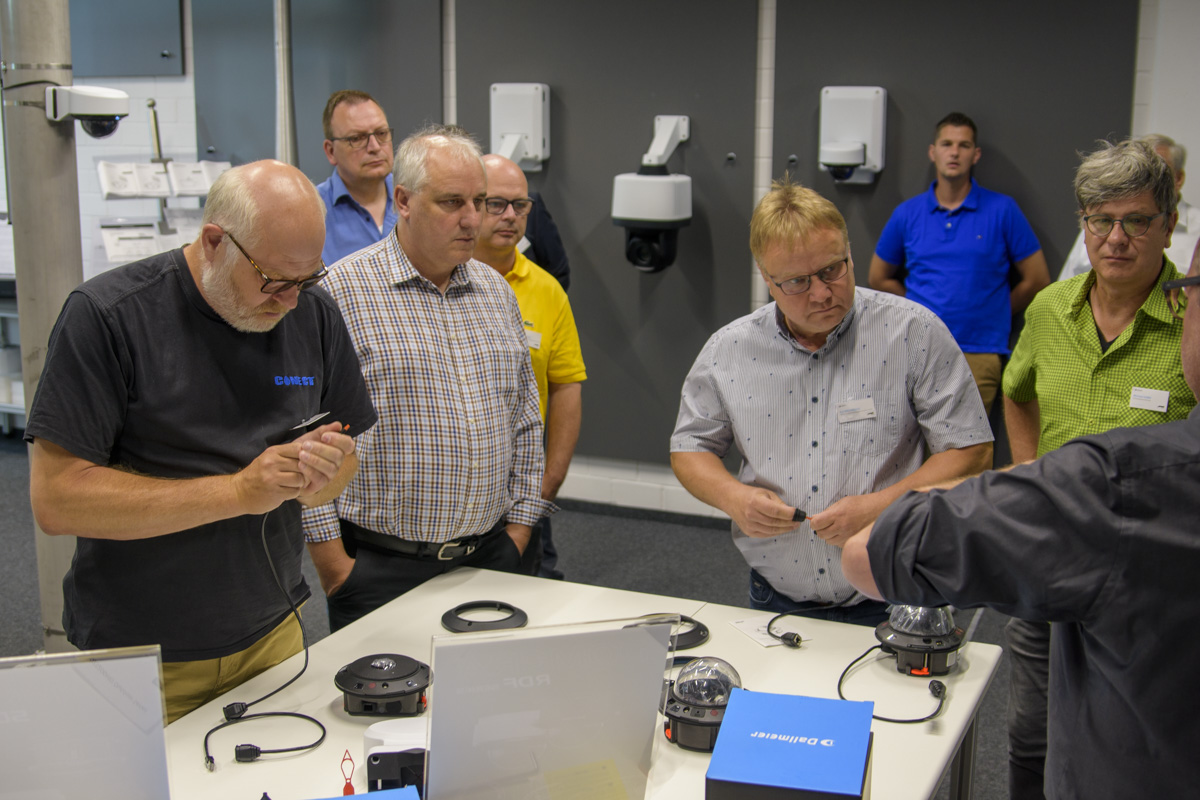 A group of people, mostly men, gather around a table displaying various electronic devices, engaged in discussion and demonstration.