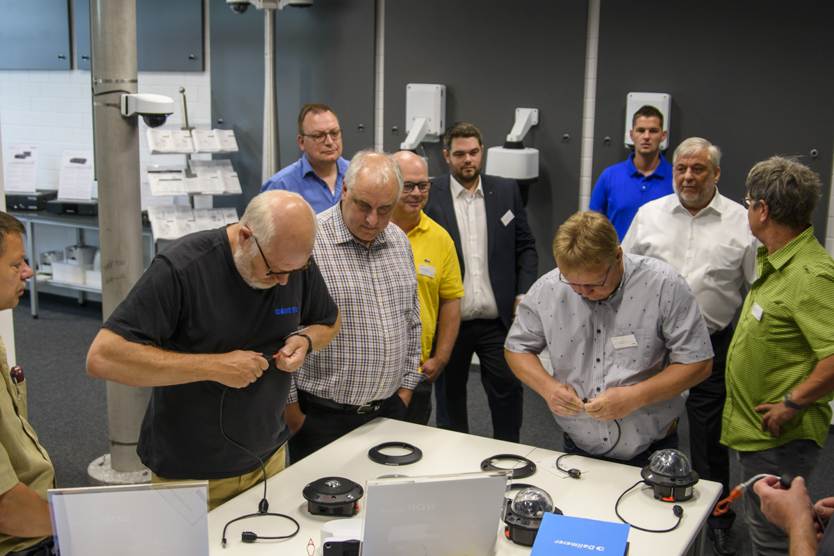 A group of men gathered around a table, focusing on hands-on electronic equipment and demonstrations in a modern workshop setting.