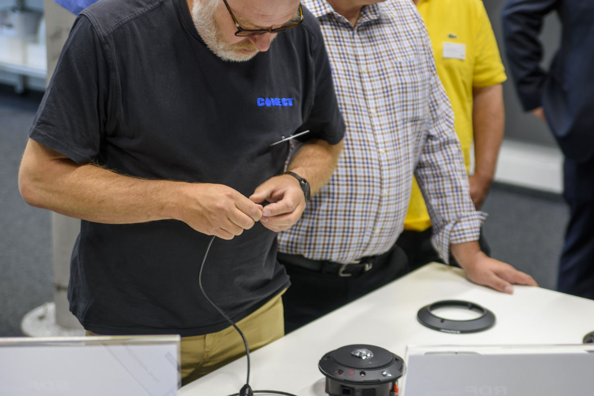 A group of people observe a man in a black shirt working on connecting a wire to a device on a table.