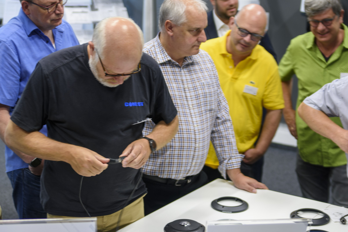 A group of six people observes a man working with a tool on a cable at a table, surrounded by various equipment and documents.