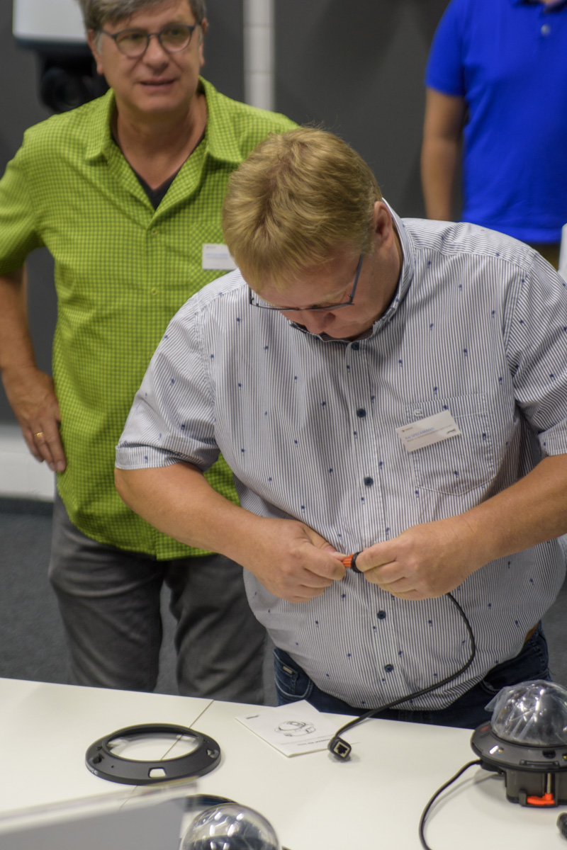 Two men are in a workshop setting. One man, in a gray shirt, is connecting wires, while the other, wearing a green checked shirt, watches attentively.