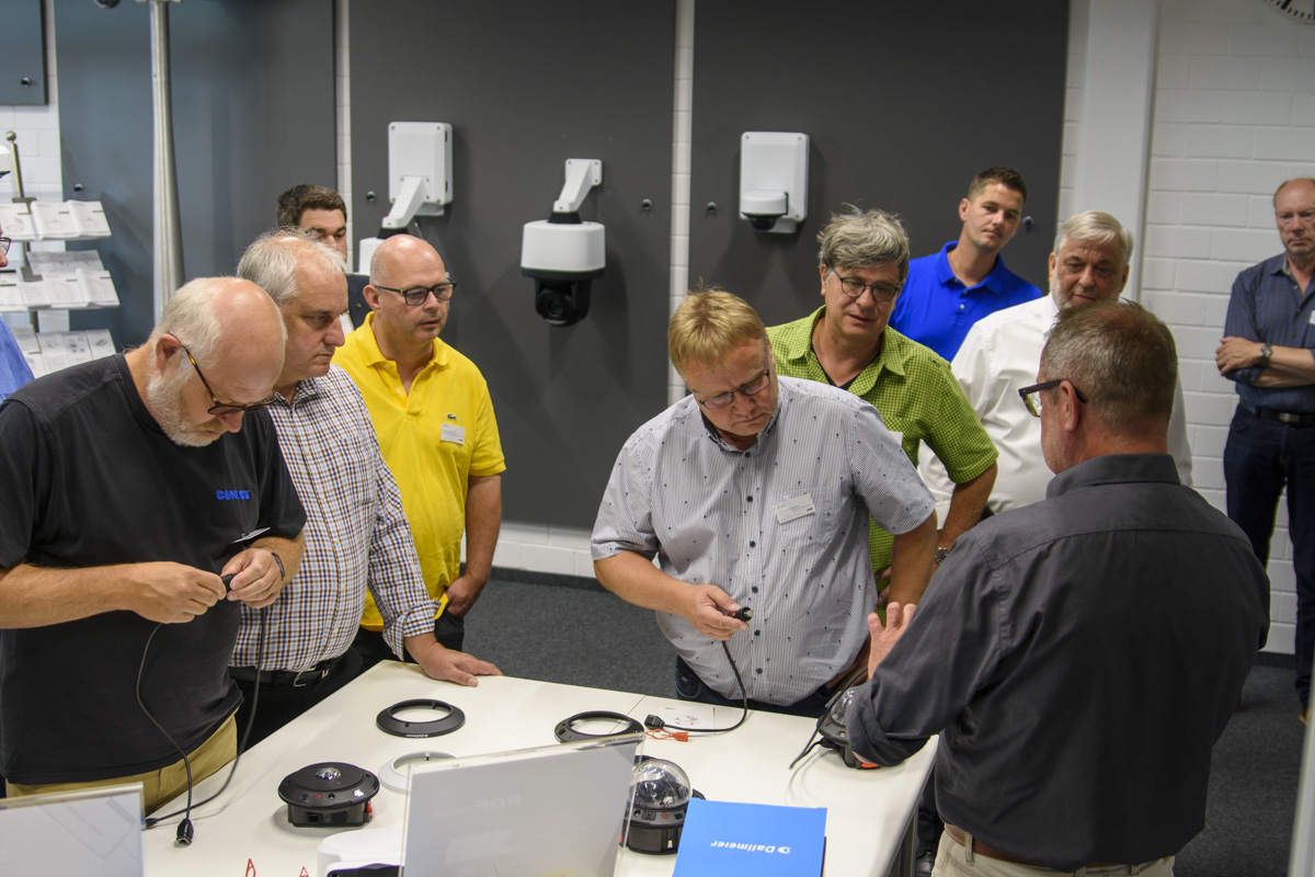 A group of professionals attentively discusses equipment at a table, with various devices and tools laid out. Some participants are taking notes or examining items.