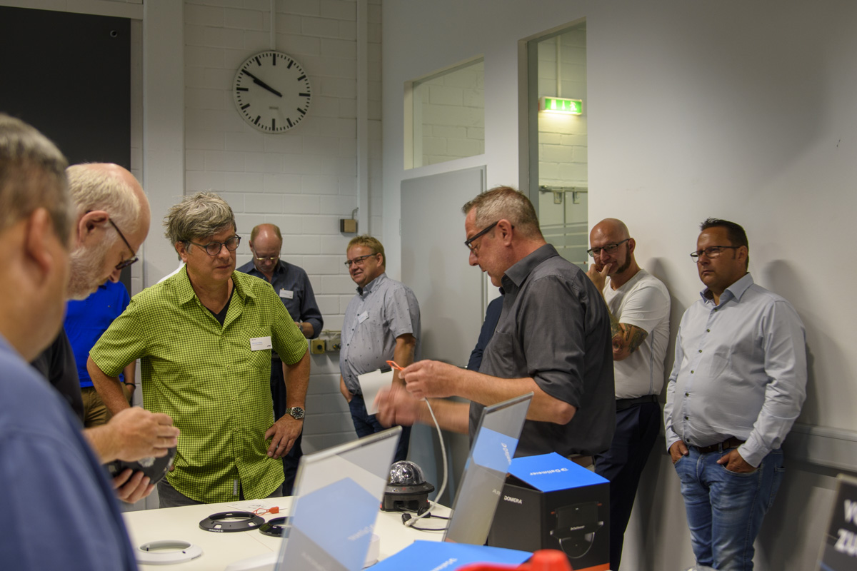 A group of people gathered around a table, engaging in discussion and observing a demonstration, with various electronics on the table and a clock visible on the wall.