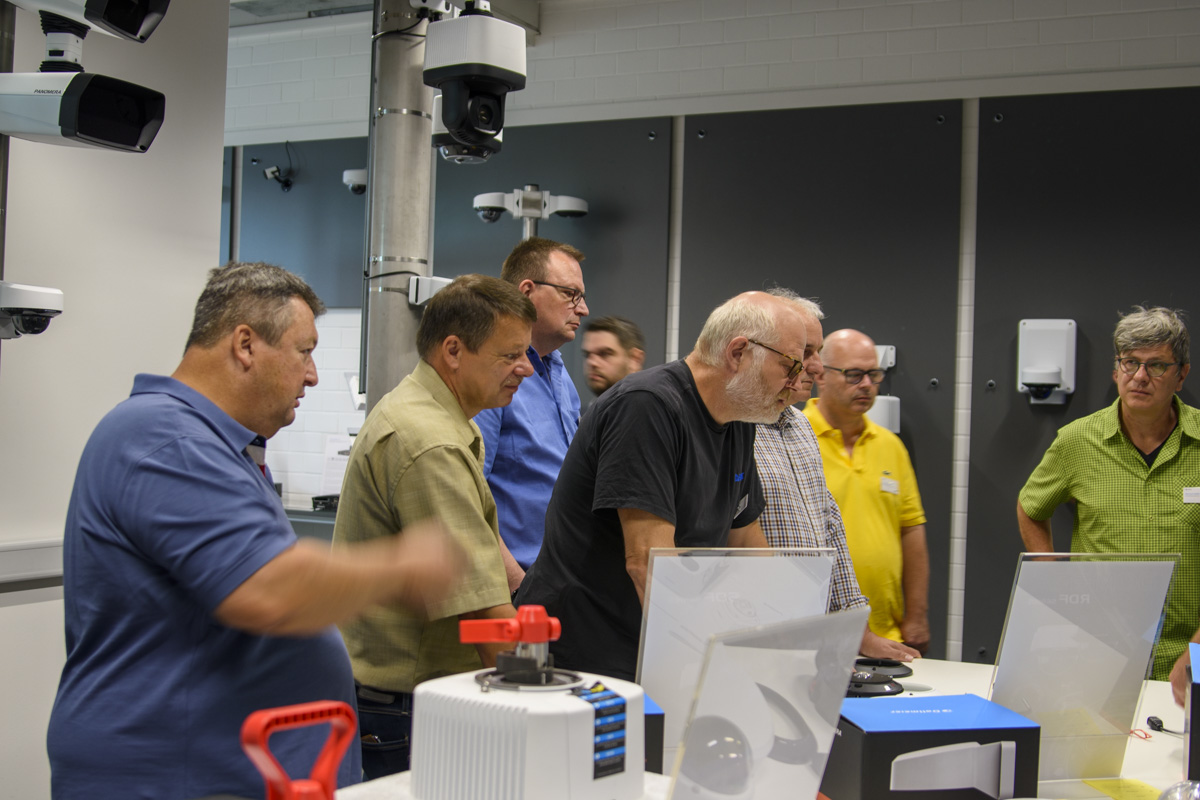 A group of six men and one woman attentively examines products on a table in a well-lit tech demonstration area, surrounded by various equipment and displays.