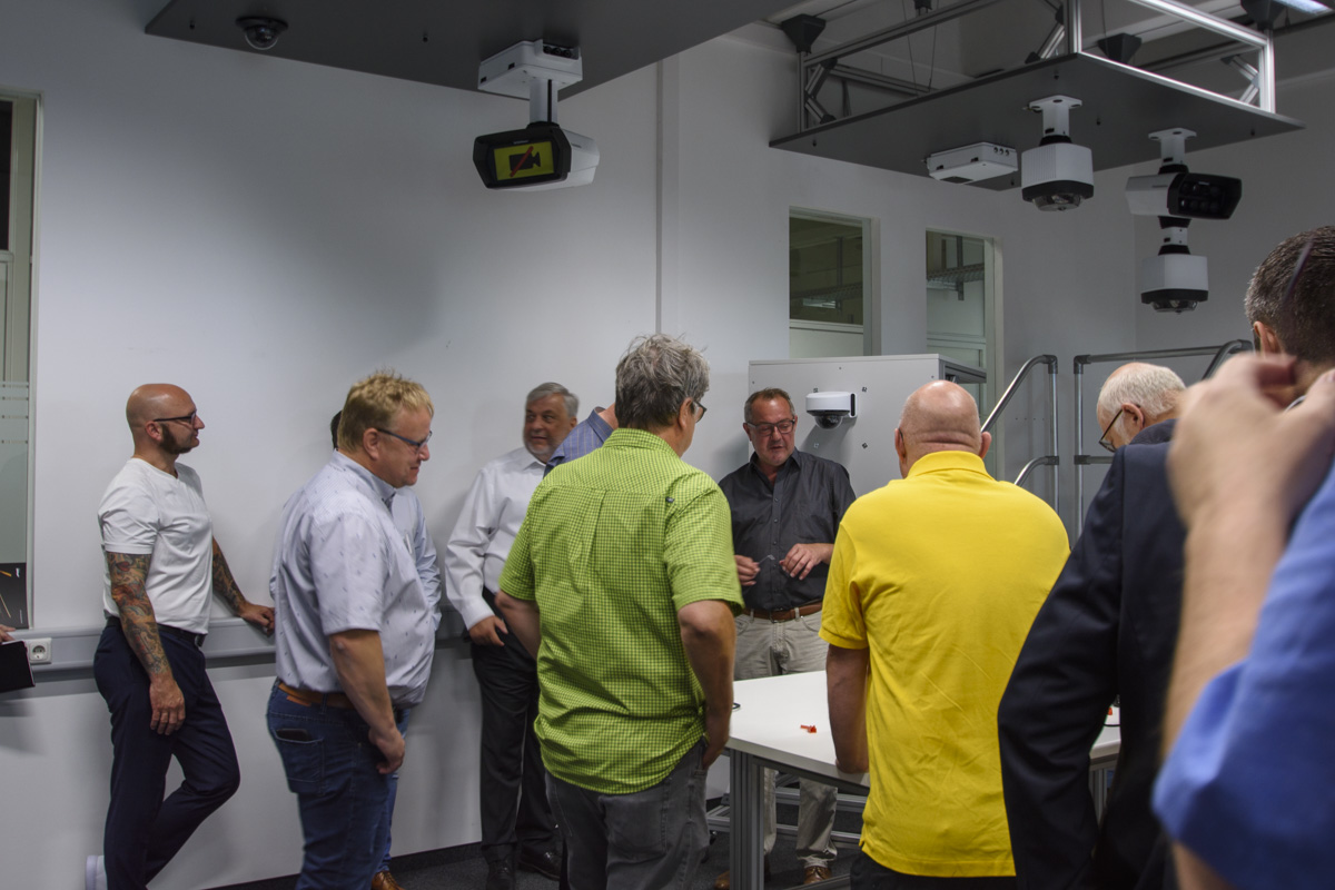 A group of eight people stands in a modern classroom, engaged in conversation. Several projectors are mounted on the ceiling. Participants wear casual attire.