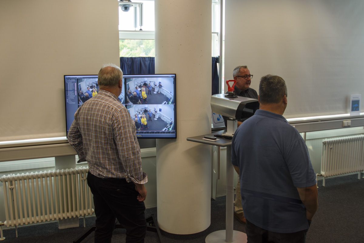 Three men stand in an indoor setting, two observing a monitor displaying multiple camera feeds, while the third speaks near a scanning device.
