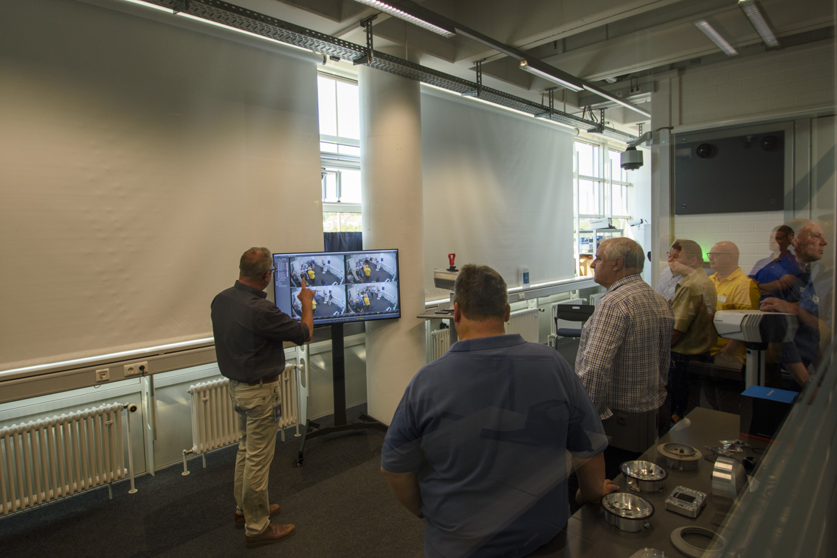 A group of people watches a presentation where a speaker interacts with a screen displaying multiple images, likely explaining a topic related to technology or training.