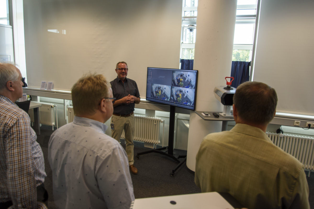 A presenter stands in front of a screen showing images while a small audience observes and listens during a presentation in a well-lit room.