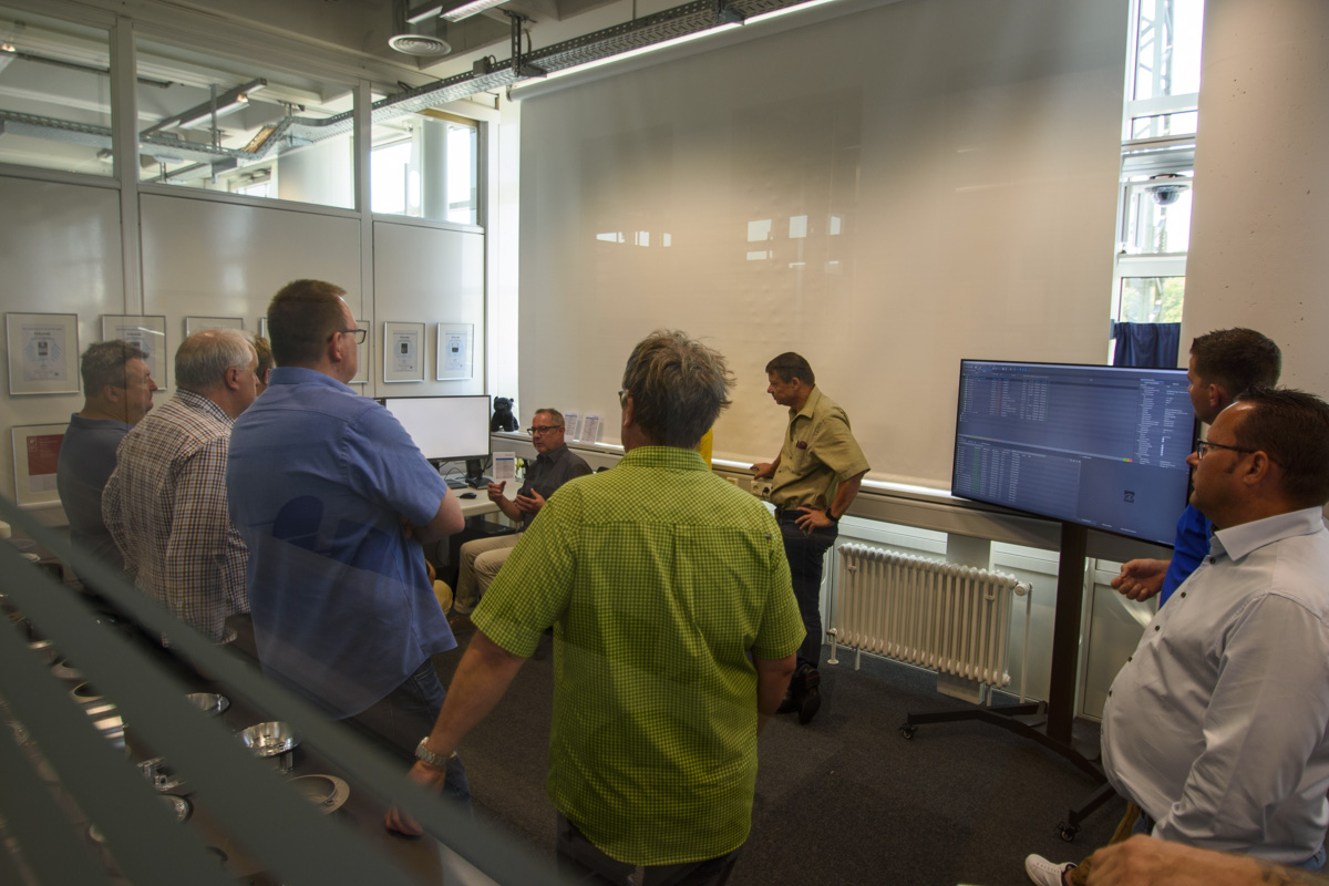 A group of eight people is gathered around a table in a meeting room, discussing information displayed on a computer screen.