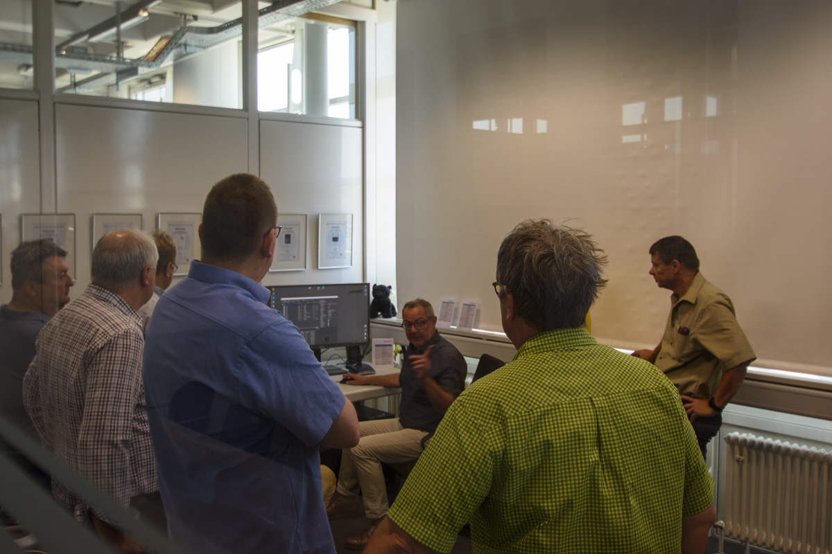 A group of men gathers in a conference room, attentively listening to a speaker who is explaining something at a computer.