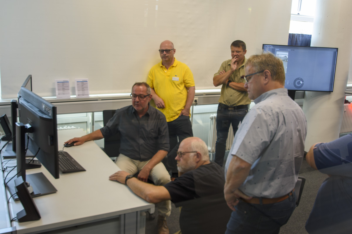 A group of five men gathered around a computer, engaged in discussion, with one man seated at the desk, while others observe and interact.