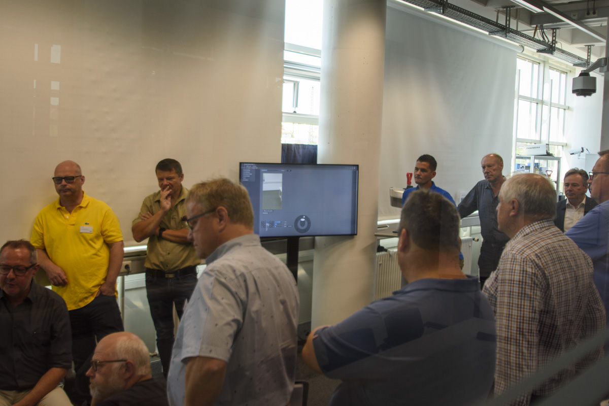 A group of men gathered in a bright modern room, some seated and others standing, facing a screen displaying content.