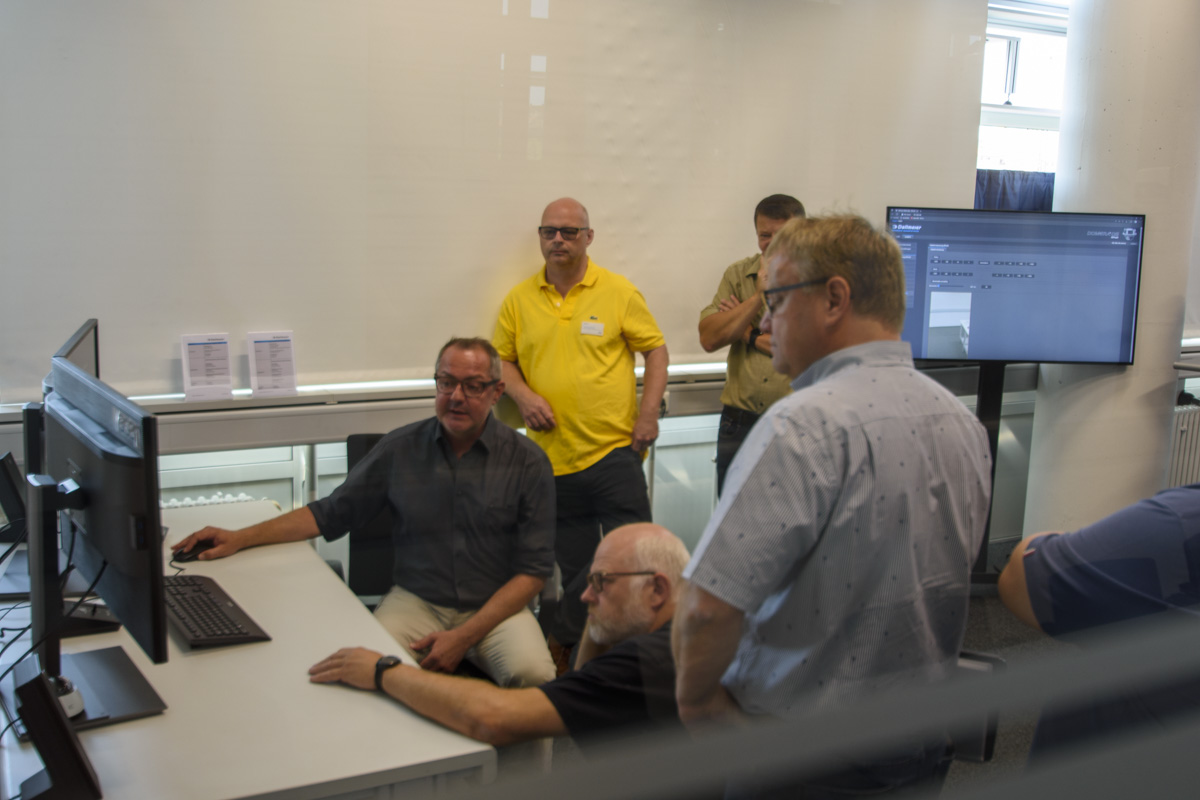A group of five men gathered around a computer, discussing something on the screen. One man is seated, while the others stand behind him, observing.
