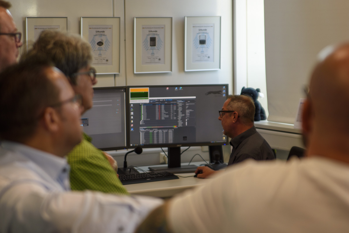 A group of people is observing a man working at a computer with dual monitors, displaying code or software, in an office setting.