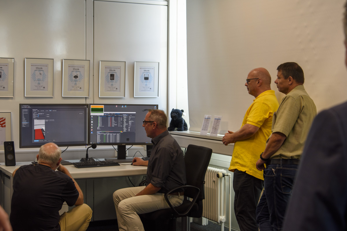 A group of four people observe a man working at a computer with dual monitors, displaying data analysis, in a modern office setting.