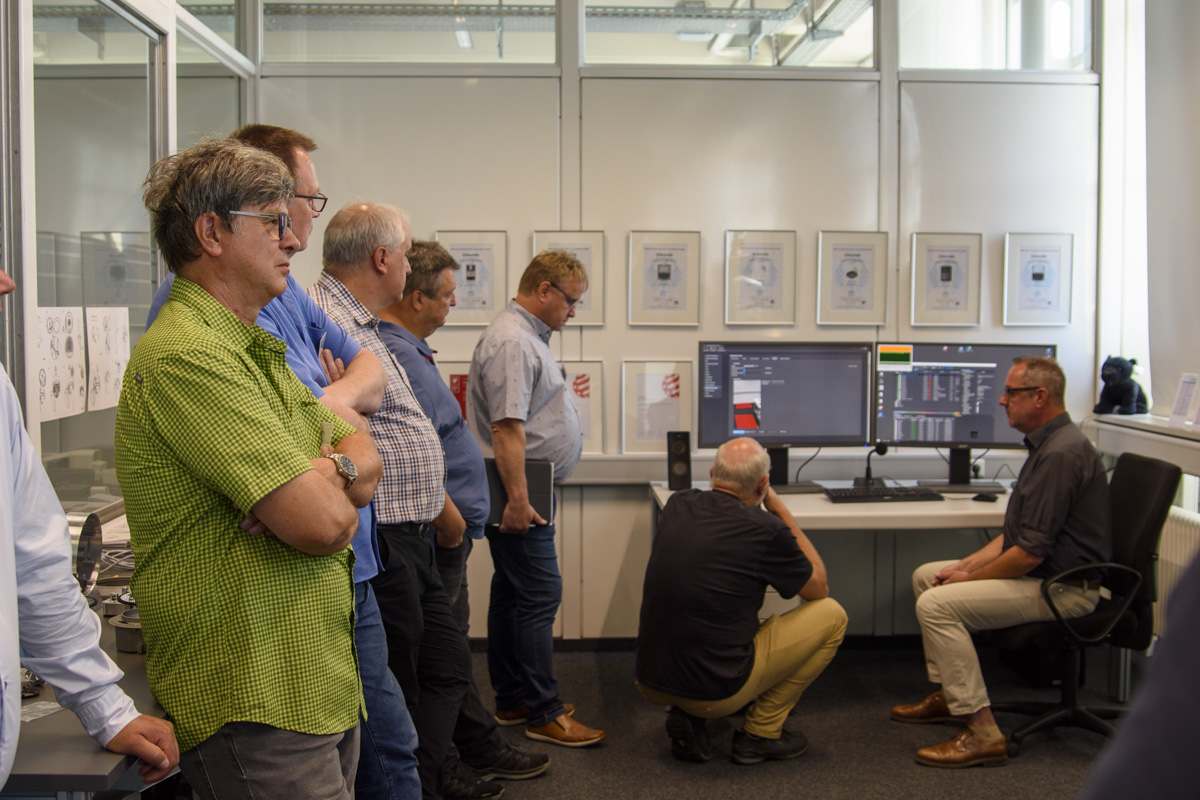 A group of eight people observes a presentation at a workstation with two monitors in an office setting. Several certificates are displayed on the walls.