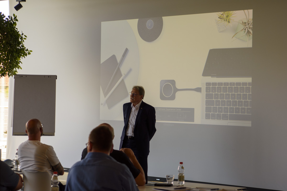 A speaker stands in front of a presentation screen displaying a minimalistic workspace design, while an audience listens attentively in a conference setting.