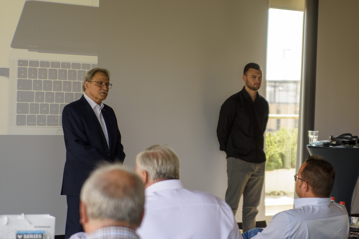 Two men are speaking in front of an audience. One man is in a suit, while the other is dressed casually. A laptop image is projected on the wall behind them.