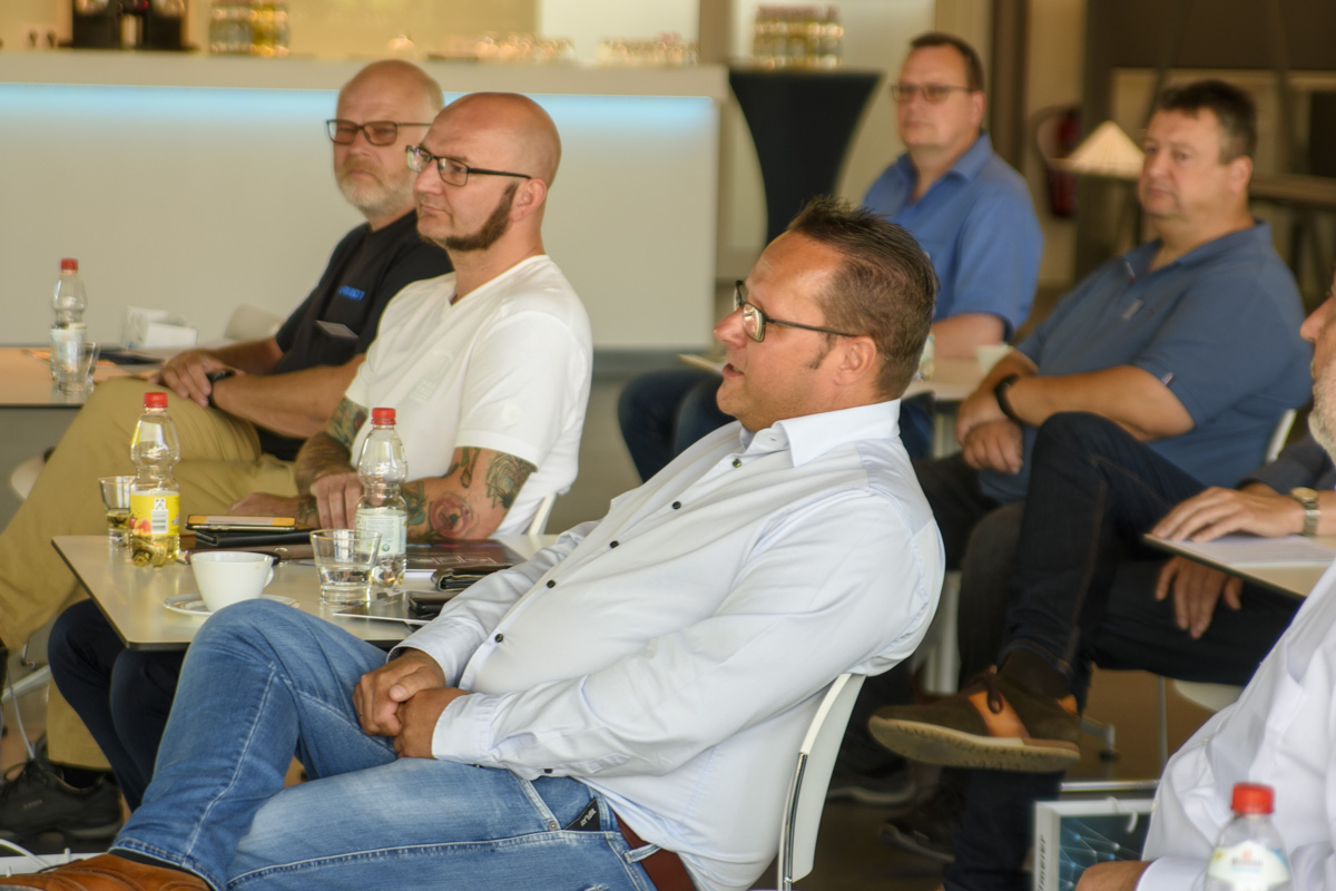 A group of men seated in a seminar setting, engaged in discussion. Some are taking notes or listening attentively. Tables in front hold drinks and papers.