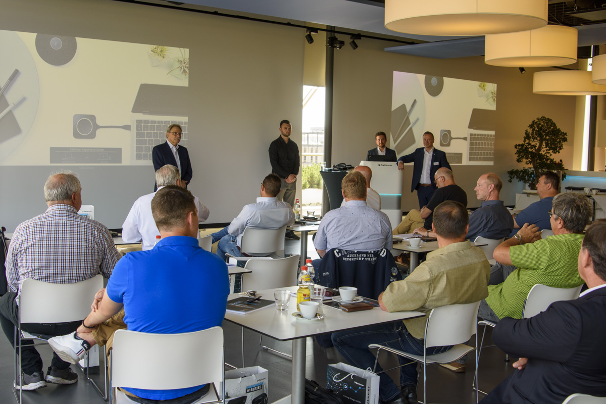 A group of people seated at tables listens to speakers presenting in a modern conference room, with large screens displaying visual content in the background.