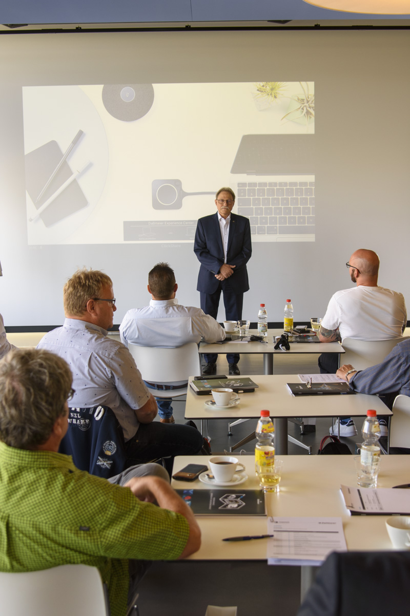 A speaker stands in front of an audience in a conference room, with a presentation projected behind. Attendees sit at tables with drinks and notebooks.