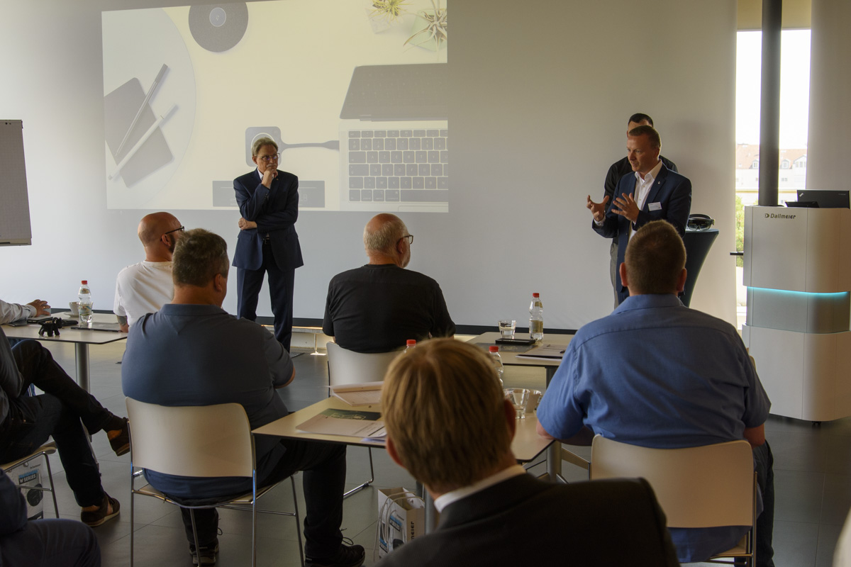 A business presentation taking place in a modern conference room, with a speaker addressing an audience seated around a table, featuring a laptop and projector screen in the background.