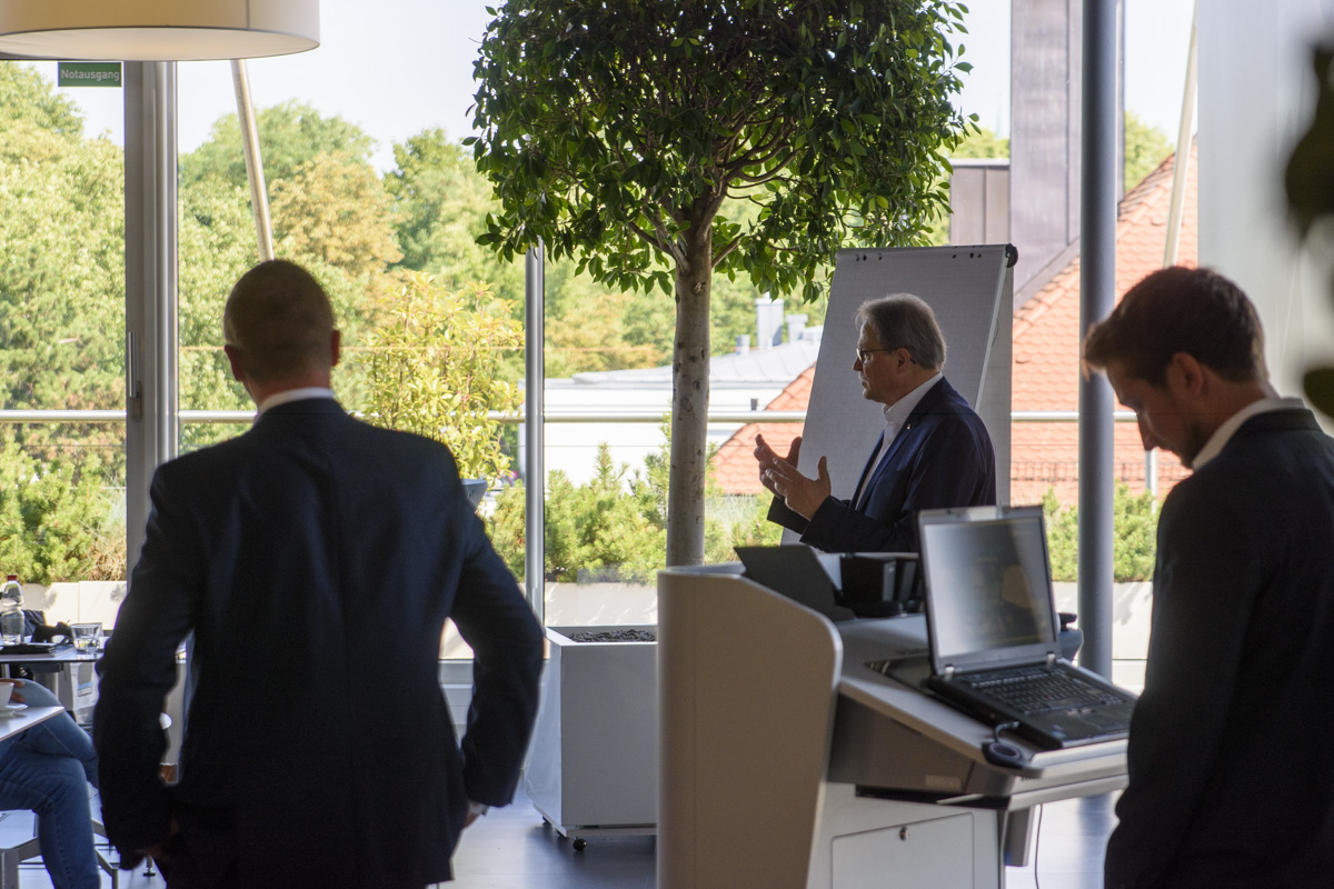 A business meeting in a modern setting, featuring a speaker gesturing at a flip chart, with attendees in suits listening attentively. Sunlight illuminates the space.