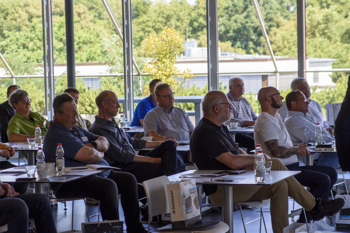 A group of people seated in a conference room, attentively listening during a presentation, with large windows showcasing greenery outside.
