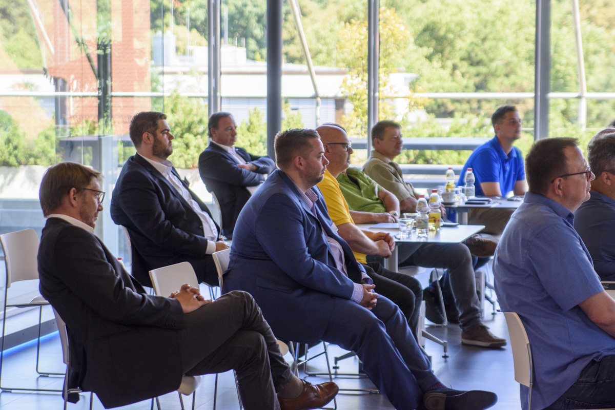 A group of attendees sitting in a modern conference room, facing forward. They are dressed in business and casual attire, with bottles of water on the tables.