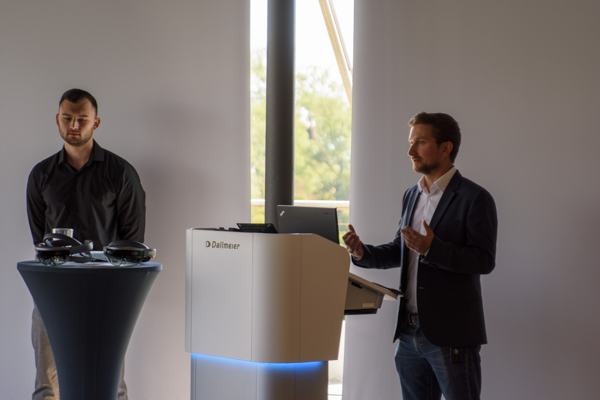 A man in a dark shirt stands next to a podium, while another man in a suit gestures as he speaks during a presentation.
